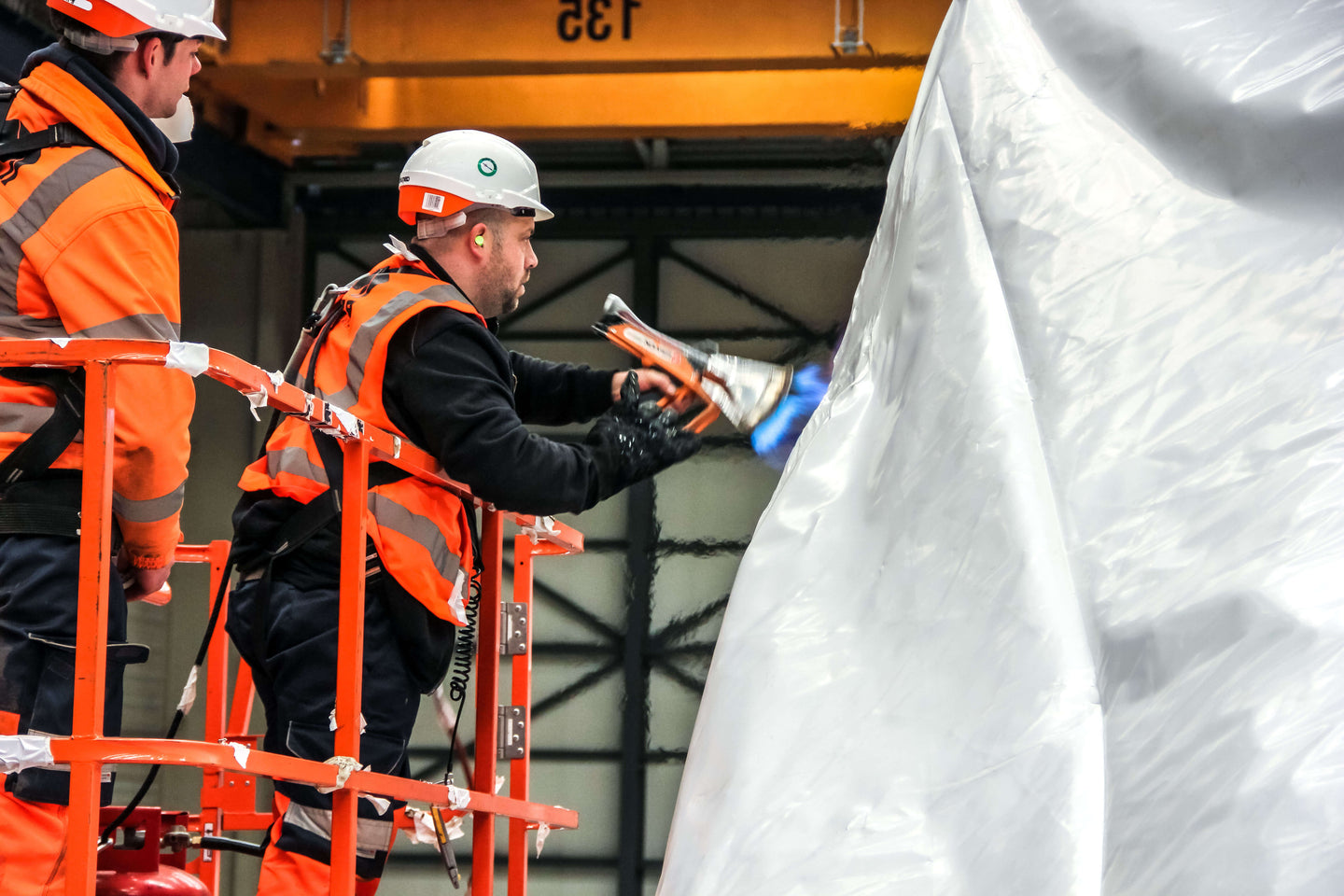 Two workers in safety gear on a lift, one using a heat tool, working on a large object.